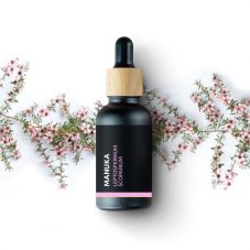 Schwarze Glasflasche mit Holzverschluss und Tropfer, enthält Bio-Manuka-Essenzielles Öl, umgeben von rosa Blüten auf weißem Hintergrund